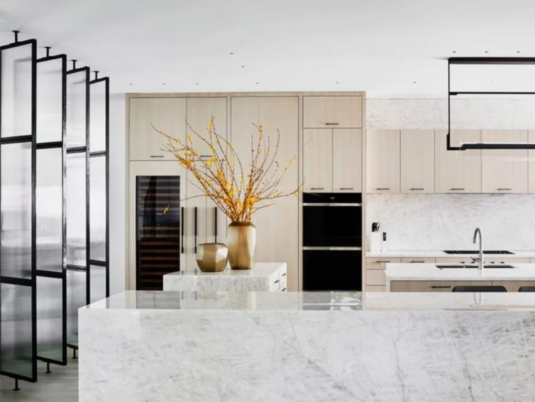 Modern kitchen with marble countertops, light wood cabinets, built-in oven, and decorative vases with yellow branches on the island